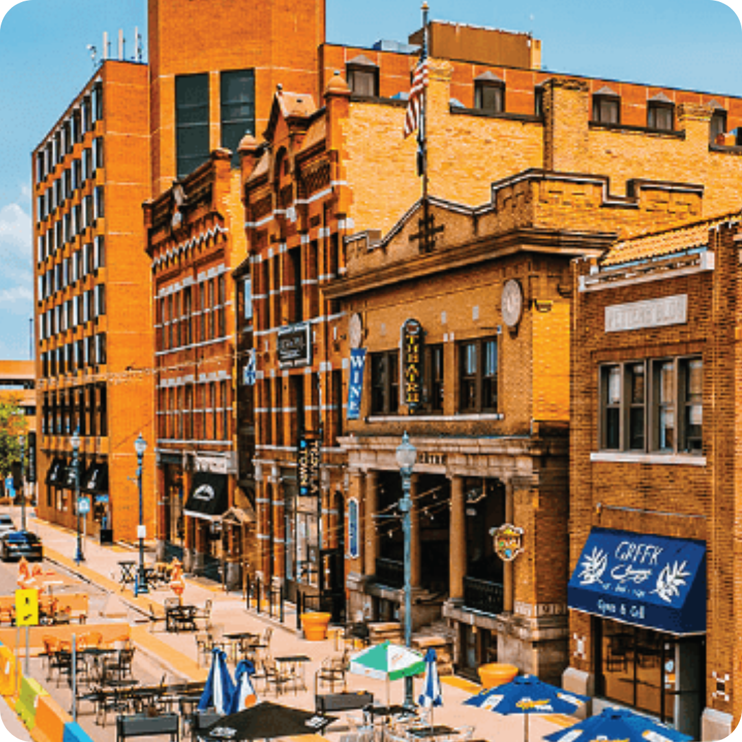 A busy urban street scene with brick buildings, outdoor dining tables, and a bright blue sky.
